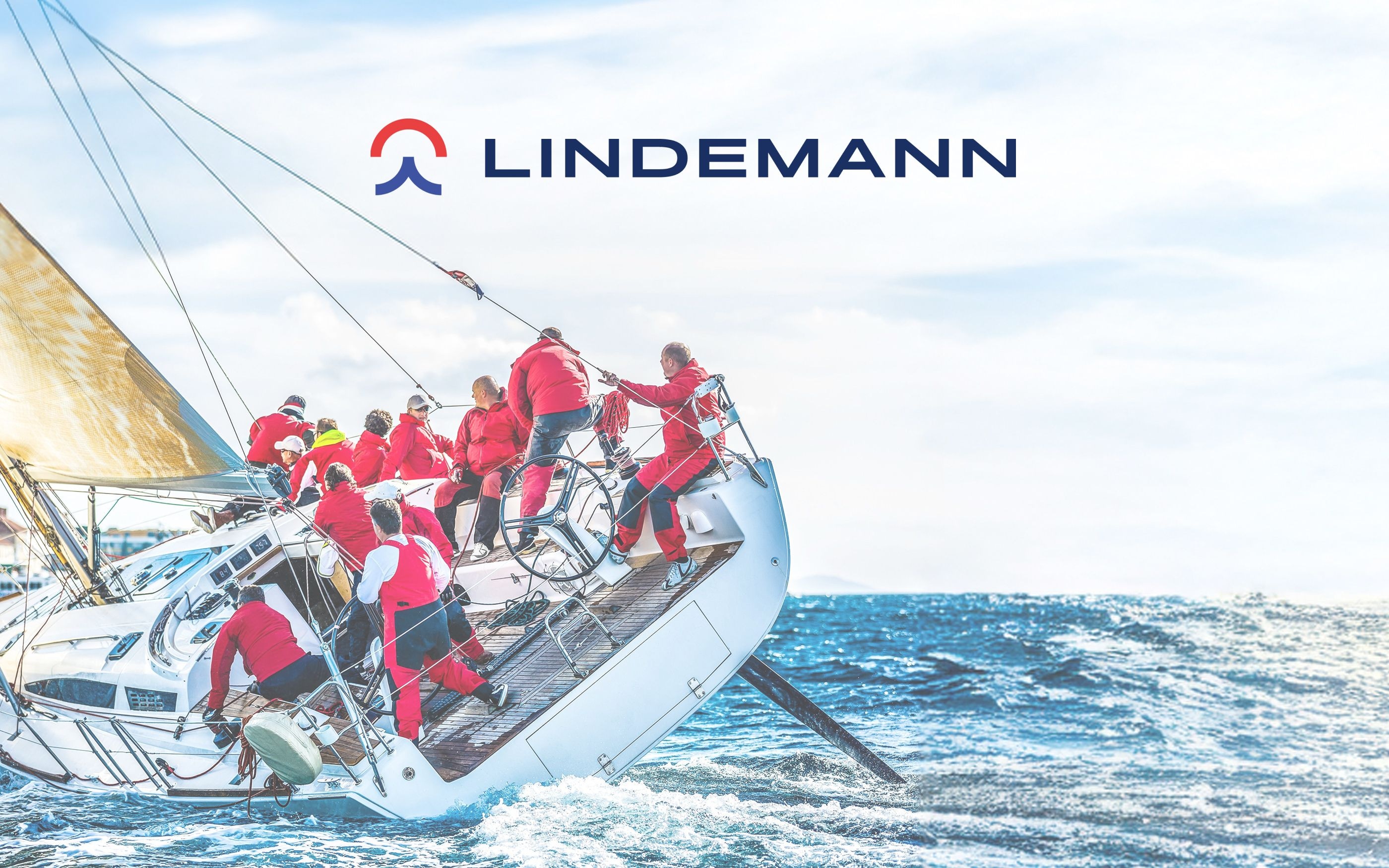 Segelboot mit Crew in roten Jacken auf stürmischem Meer, stark geneigt nach Steuerbord. Im Hintergrund blauer Himmel mit Wolken. Oben das Lindemann-Logo in Blau und Rot.