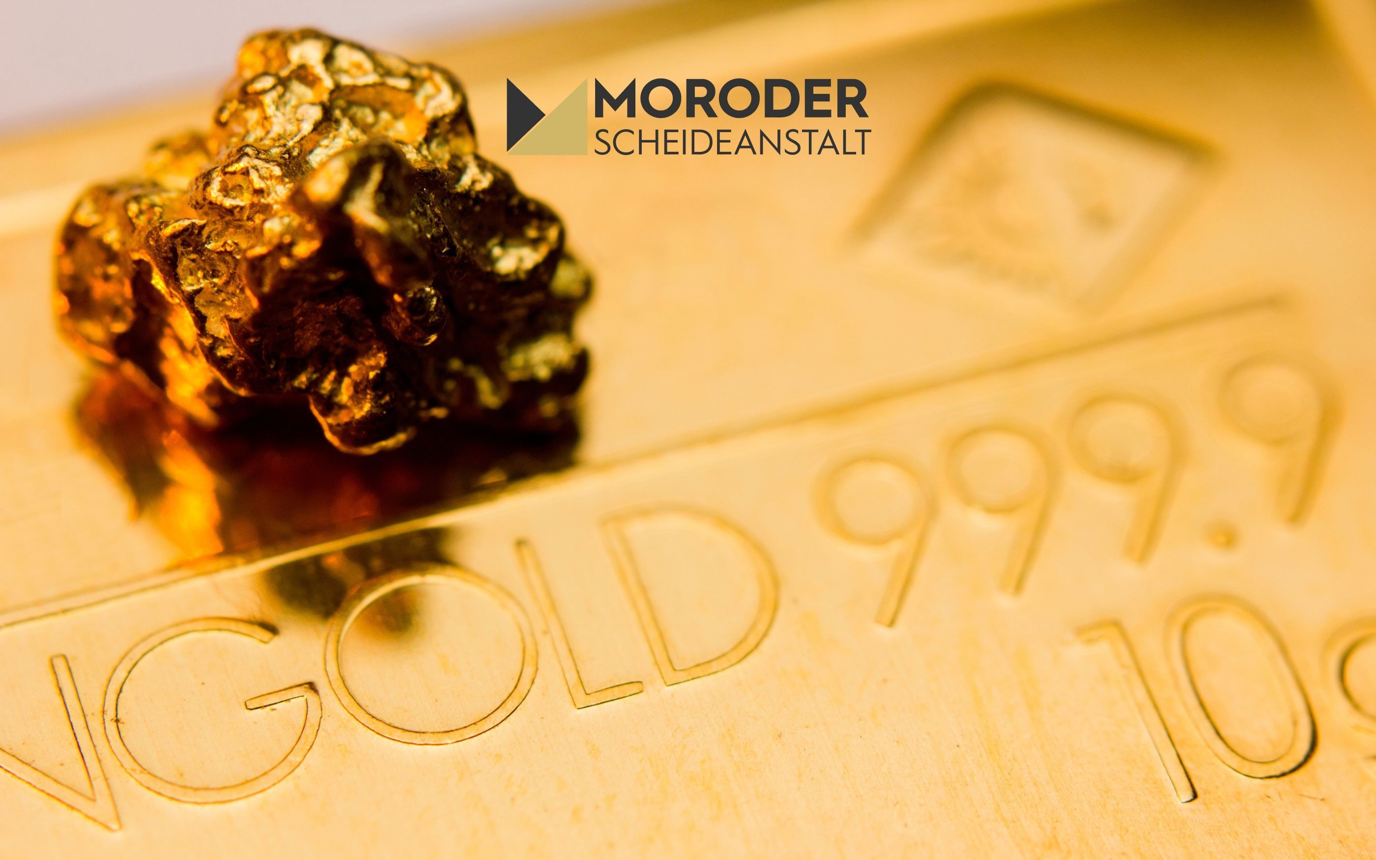 Nahaufnahme eines Goldbarrens mit eingeprägter Aufschrift ‚GOLD 999.9 10g‘ und einem kleinen Goldnugget darauf. Im Hintergrund das Logo ‚Moroder Scheideanstalt‘.